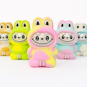 May include: A group of brightly coloured figurines, each resembling a creature in a hooded costume. The figures have smiling faces and are predominantly yellow, pink, green, and blue. The design is cartoonish and playful.