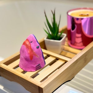 Handmade pink shark bath bomb resting over the bathtub. The bath bomb features a cute, hand-painted shark face with vibrant pink colour, perfect for a fun and fizzy bath time experience.