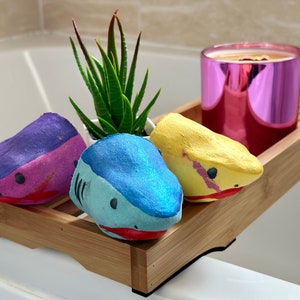 Shark Bath Bomb Gift Set displayed over a bathtub. The set includes three handmade, hand-painted shark-shaped bath bombs in blue, pink, and yellow. Each bath bomb has a friendly shark face and vibrant colours, perfect for kids and bath lovers. A fun and skin-nourishing bath experience in a beautifully crafted gift set.