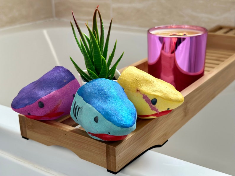 Shark Bath Bomb Gift Set displayed over a bathtub. The set includes three handmade, hand-painted shark-shaped bath bombs in blue, pink, and yellow. Each bath bomb has a friendly shark face and vibrant colours, perfect for kids and bath lovers. A fun and skin-nourishing bath experience in a beautifully crafted gift set.