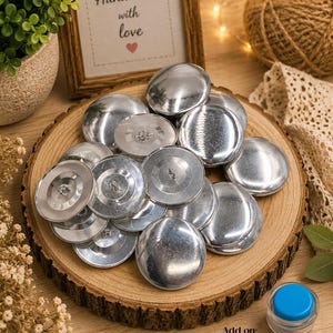 May include: A collection of silver-colored, domed buttons, size 60 (1 1/2 inches), displayed on a wooden slice. Some buttons have a flat back with a loop. A small container with a blue lid and the text "Add on: Assembly Tool" is visible.