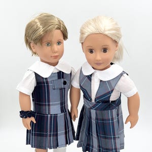 May include: Two dolls wearing matching plaid school uniforms with white shirts and collars. The dolls have blonde hair and are wearing black shoes with gold buckles.