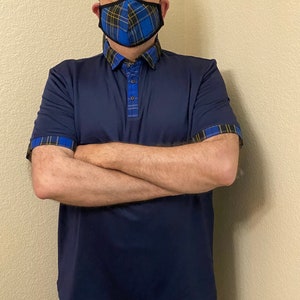 May include: A man wearing a navy blue polo shirt with plaid trim on the collar and sleeves. He is wearing a blue and yellow plaid face mask.