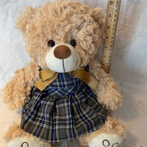 May include: A beige plush teddy bear wearing a blue and yellow plaid dress with a gold ribbon. The bear is sitting on a white fluffy surface. The bear is approximately 11 inches tall.