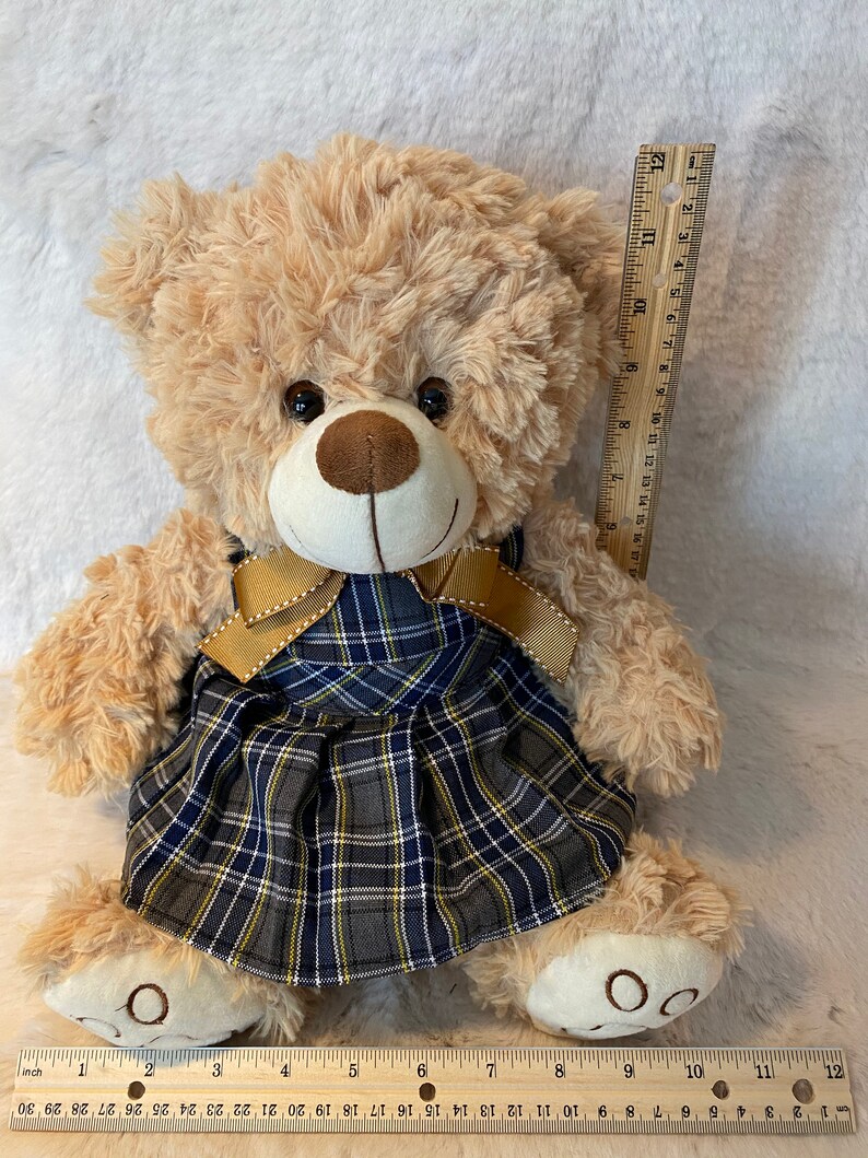 May include: A beige plush teddy bear wearing a blue and yellow plaid dress with a gold ribbon. The bear is sitting on a white fluffy surface. The bear is approximately 11 inches tall.