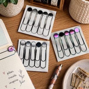 May include: An assortment of stationery items on a wooden surface. Includes silver, black, and iridescent paper clips, a patterned pen, a notebook with the words "plan create inspire", and decorative washi tape.