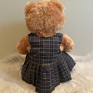 May include: A brown teddy bear wearing a plaid dress with a pleated skirt.