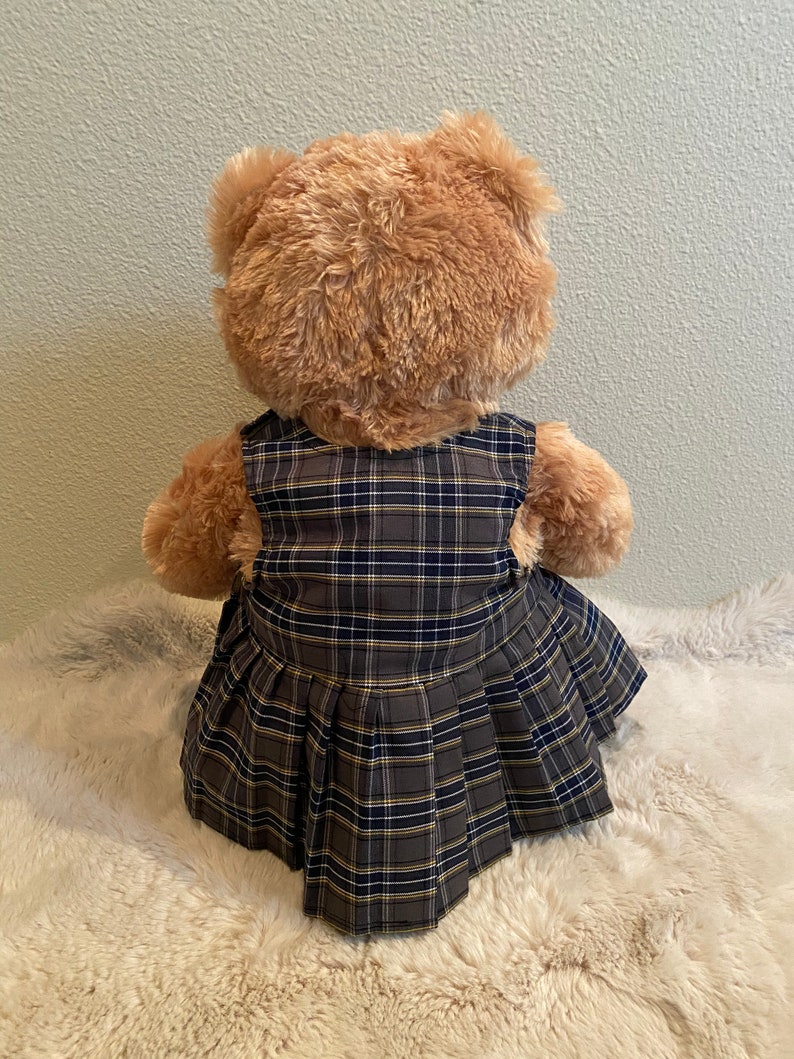 May include: A brown teddy bear wearing a plaid dress with a pleated skirt.