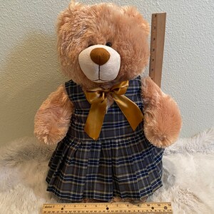May include: A brown teddy bear wearing a plaid dress with a gold bow. The dress has a black, white and yellow plaid pattern.
