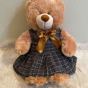 May include: A brown teddy bear wearing a plaid dress with a gold bow. The dress has a navy blue, yellow, and white plaid pattern.