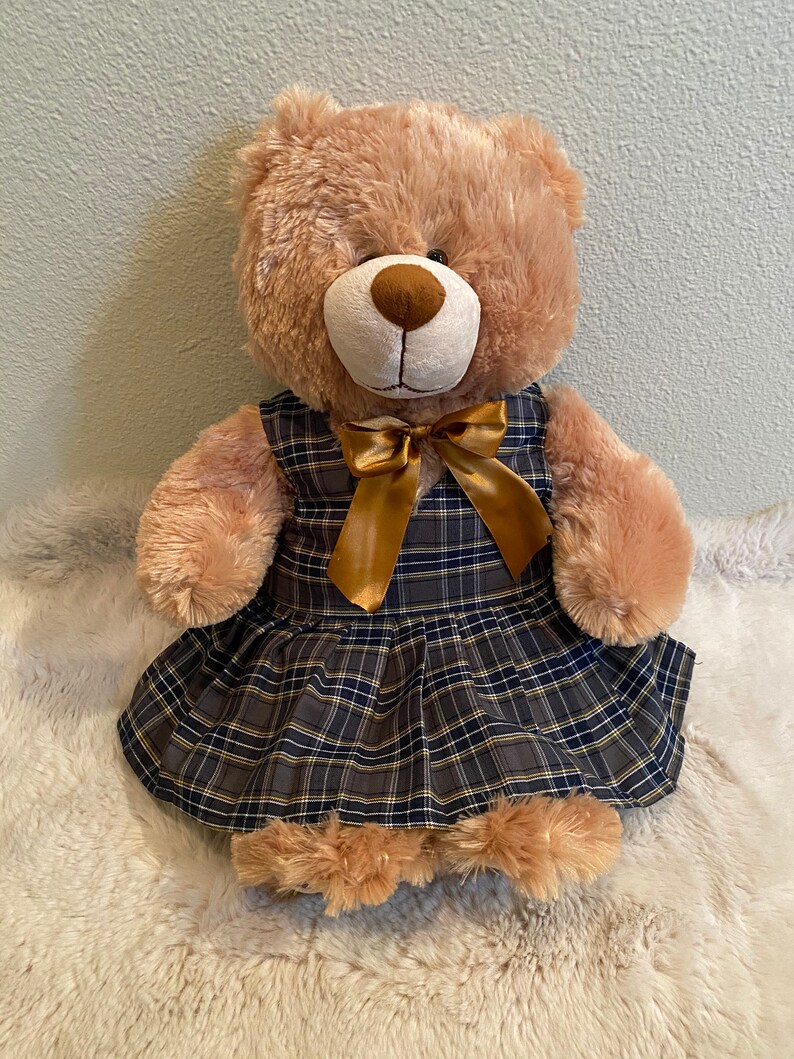 May include: A brown teddy bear wearing a plaid dress with a gold bow. The dress has a navy blue, yellow, and white plaid pattern.