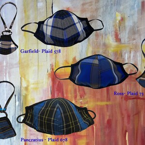 Op de afbeelding: Vier verschillende geruite mondmaskers met zwarte elastieken en een bijpassend geruit koord. De maskers zijn blauw, zwart en wit met verschillende ruitpatronen. De koorden zijn met een kleine metalen ring aan de maskers bevestigd. De maskers zijn gelabeld met "Garfield - Plaid 578", "Ross - Plaid 73" en "Pancratius - Plaid 678".