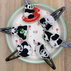 Op de afbeelding: Zes zwart-witte pandabeer haarclips op een wit en groen bord met een ster- en hartpatroon. De pandaberen zijn in verschillende poses, waaronder zittend op een schommelpaard, een bamboescheut vasthoudend en slapen.