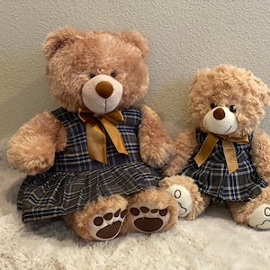 May include: Two plush teddy bears wearing plaid dresses with gold bows. The larger bear is sitting with its legs crossed, while the smaller bear is sitting with its legs straight out in front of it.
