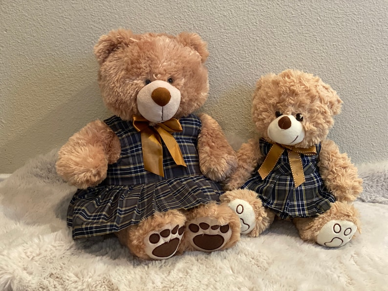 May include: Two plush teddy bears wearing plaid dresses with gold bows. The larger bear is sitting with its legs crossed, while the smaller bear is sitting with its legs straight out in front of it.