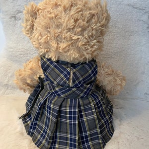 May include: A beige teddy bear wearing a grey, blue and yellow plaid dress with a pleated skirt.