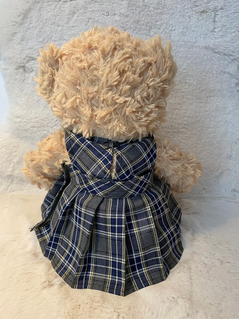 May include: A beige teddy bear wearing a grey, blue and yellow plaid dress with a pleated skirt.