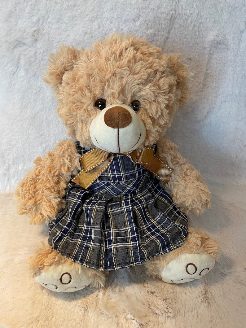 May include: A plush teddy bear wearing a plaid dress with a brown bow. The bear has light brown fur and a white face with brown eyes.