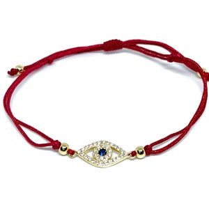 May include: Red string bracelet with a gold evil eye charm. The charm is decorated with clear and blue crystals.