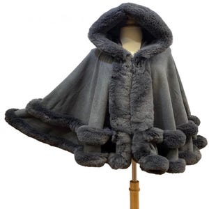 May include: A dark gray hooded cape featuring thick, plush faux fur trim along the hood, the front opening, and the scalloped hemline. The garment has a smooth, solid-colored fabric body that drapes outward from the shoulders in a wide, flared silhouette.