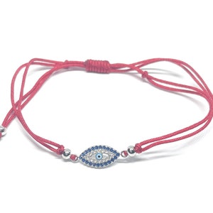 May include: Red string bracelet with a silver evil eye charm. The charm is decorated with blue and white stones.