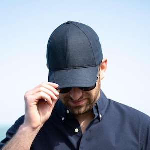 May include: A person wearing a dark blue button-down shirt and a dark blue baseball cap. The person is standing in front of a body of water.