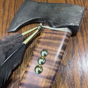 May include: A handmade axe with a wooden handle and a metal head. The handle is decorated with brass accents and a leather wrap. The axe is resting on a wooden surface.