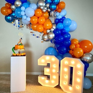 Könnte beinhalten: Eine Geburtstagsdekoration mit blauen, orangen und silbernen Luftballons, die in einer Kaskade angeordnet sind. Die Ballons hängen an einer Wand und sind mit einem Kuchen und den Zahlen 30 in weißen Lichtern verziert.