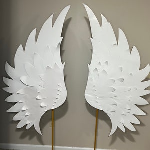 May include: A pair of large white paper angel wings on gold metal stands. The wings are made of multiple layers of paper, creating a 3D effect.