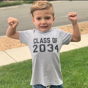 May include: A young boy wearing a gray t-shirt with the text "CLASS OF 2034" printed on it. He is wearing blue jeans and has a short haircut.