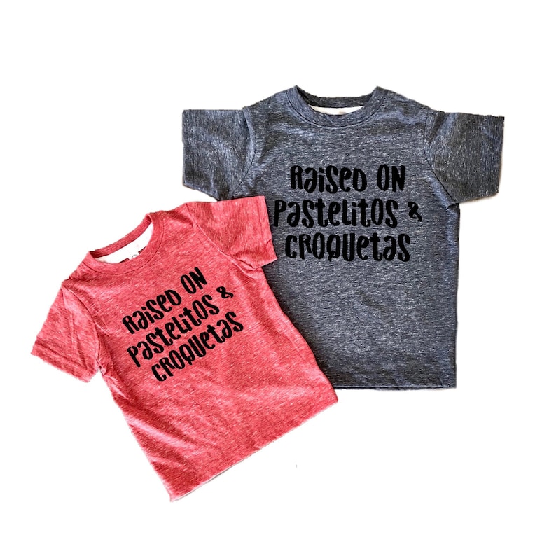 Op de afbeelding: Twee t-shirts voor peuters met de tekst "raised on pastelitos & croquetas" in het zwart. E&eacute;n shirt is rood en de andere is grijs.