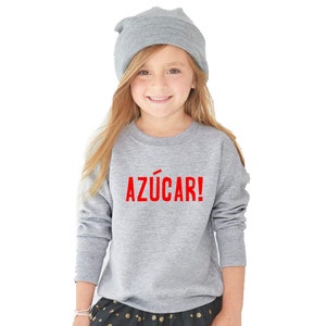 May include: A young girl wearing a gray sweatshirt with the word "AZ&Uacute;CAR!" printed in red on the front. She is also wearing a gray beanie and a black and white polka dot skirt.