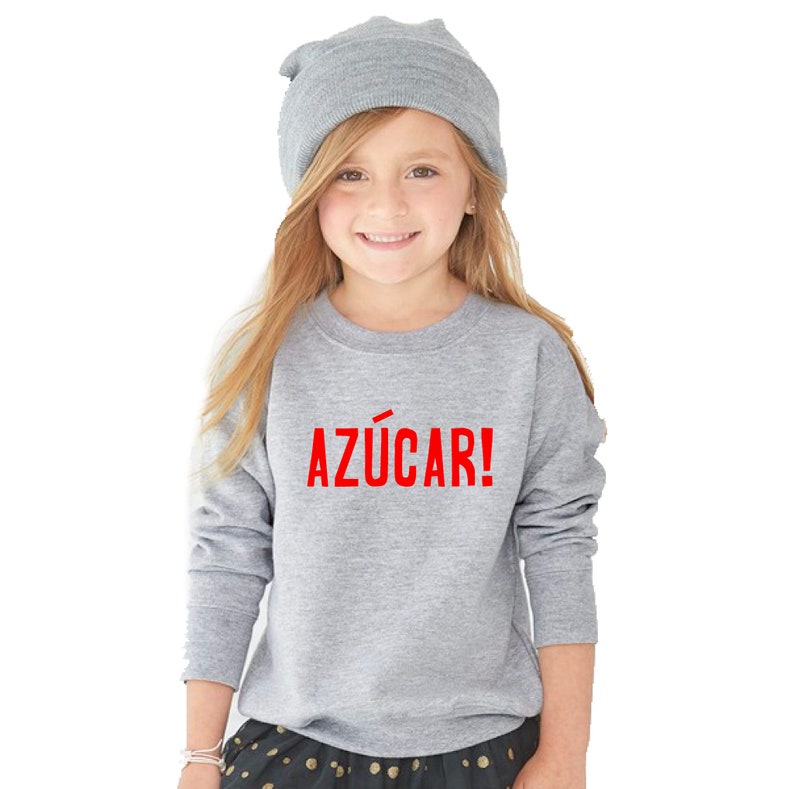 May include: A young girl wearing a gray sweatshirt with the word "AZ&Uacute;CAR!" printed in red on the front. She is also wearing a gray beanie and a black and white polka dot skirt.