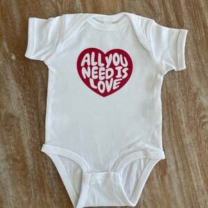 May include: White baby onesie with a red heart graphic that says "All you need is love" in white lettering.
