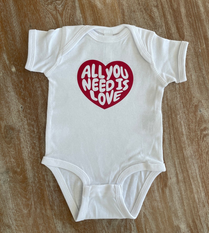 May include: White baby onesie with a red heart graphic that says "All you need is love" in white lettering.