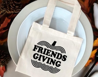 以下が含まれることがあります： 黒いハンドルと「FRIENDS GIVING」というレトロなフォントのテキストが黒いカボチャの輪郭の中に書かれた白いキャンバス地のトートバッグです。