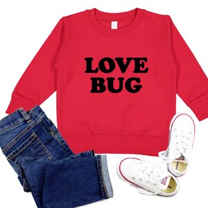 May include: Red sweatshirt with black text that says "LOVE BUG". The sweatshirt is paired with blue jeans and white sneakers.