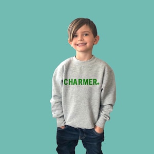 Puede incluir: Una persona joven lleva una sudadera gris con la palabra "CHARMER" impresa en letras verdes. La persona lleva pantalones vaqueros azules y tiene el pelo corto y castaño.