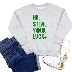 Pode incluir: Um moletom cinza claro com o texto verde "MR. STEAL YOUR LUCK" e um trevo verde. A imagem também mostra jeans azuis e tênis de lona brancos com detalhes em vermelho. Os itens são dispostos sobre um fundo branco.