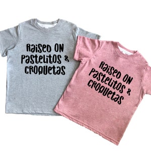 Op de afbeelding: Twee T-shirts met de tekst "Raised on pastelitos & croquetas" in zwarte letters. Een shirt is grijs en de andere is roze.