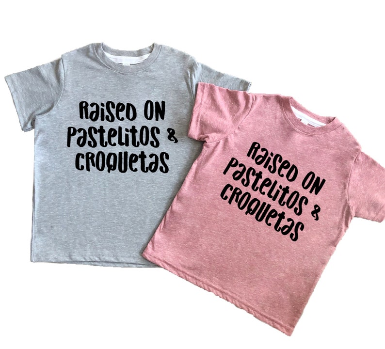 Op de afbeelding: Twee T-shirts met de tekst "Raised on pastelitos & croquetas" in zwarte letters. Een shirt is grijs en de andere is roze.