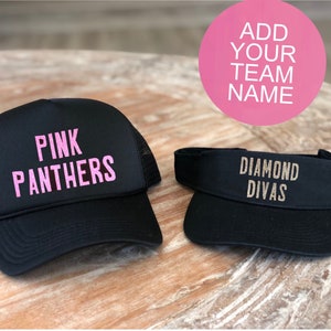 Può includere: Due cappellini da baseball neri con nomi di squadra diversi. Il primo cappellino è uno stile camionista con una rete rosa sul retro e le parole "PINK PANTHERS" in lettere rosa. Il secondo cappellino è una visiera con le parole "DIAMOND DIVAS" in lettere dorate glitter.
