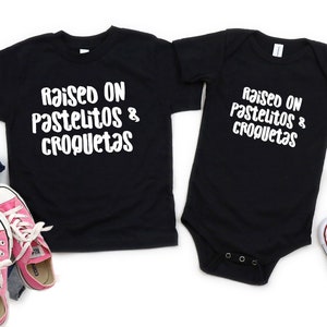 Op de afbeelding: Twee zwarte T-shirts met de tekst "Raised on pastelitos & croquetas" in wit. Het grotere shirt is een kindermaat en het kleinere shirt is een baby-romper.