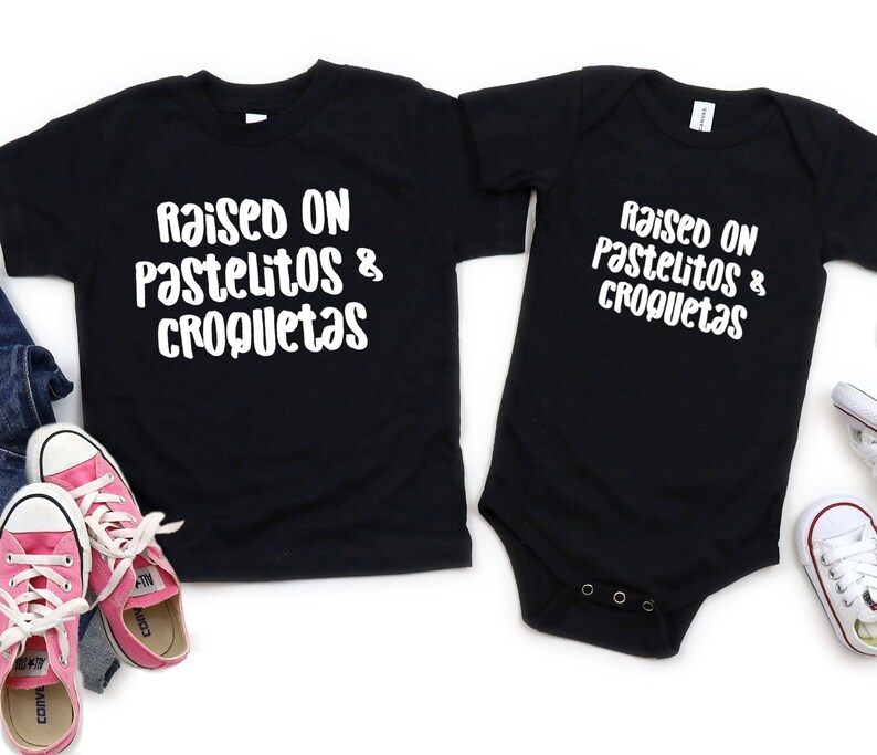 Op de afbeelding: Twee zwarte T-shirts met de tekst "Raised on pastelitos & croquetas" in wit. Het grotere shirt is een kindermaat en het kleinere shirt is een baby-romper.