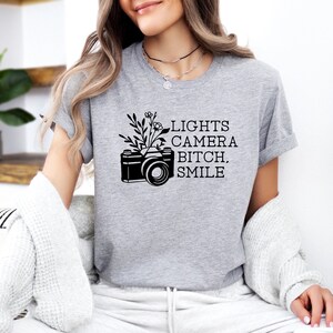 Op de afbeelding: Een grijs T-shirt met een zwarte camera en een bloemenontwerp. De tekst "LIGHTS CAMERA BITCH, SMILE" staat op het T-shirt gedrukt.