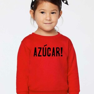 May include: A red sweatshirt with the text "AZ&Uacute;CAR!" in black lettering. A young girl is wearing the sweatshirt and has a black and white polka dot headband in her hair.