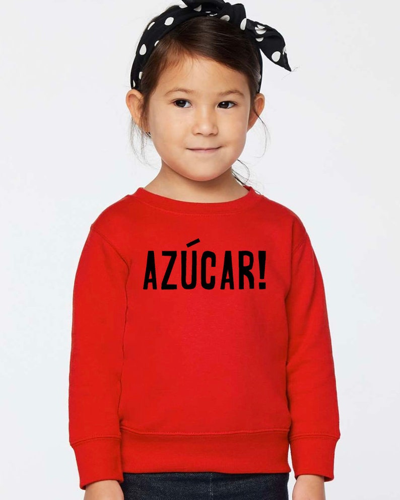 May include: A red sweatshirt with the text "AZ&Uacute;CAR!" in black lettering. A young girl is wearing the sweatshirt and has a black and white polka dot headband in her hair.