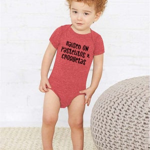 Op de afbeelding: Een rood babypakje met de tekst "Raised on Pastelitos & Croquetas" in zwart gedrukt.