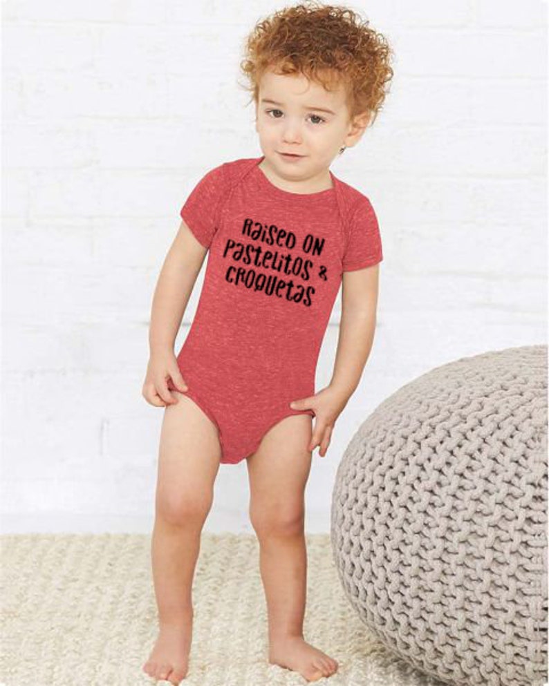 Op de afbeelding: Een rood babypakje met de tekst "Raised on Pastelitos & Croquetas" in zwart gedrukt.