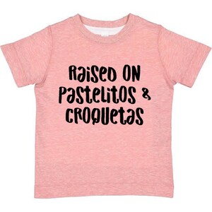 Op de afbeelding: Roze T-shirt met korte mouwen met de tekst "Raised on Pastelitos & Croquetas" in zwart.
